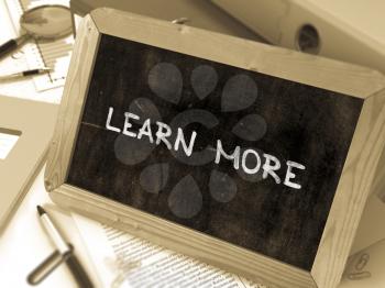 Learn More Handwritten on Chalkboard. Composition with Small Chalkboard on Background of Working Table with Ring Binders, Office Supplies, Reports. Blurred Background. Toned Image. 3d Render.