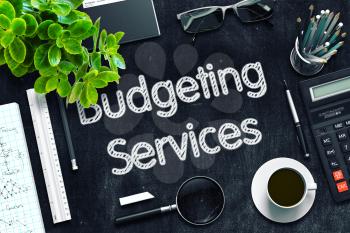 Budgeting Services Handwritten on Black Chalkboard. Top View of Black Office Desk with a Lot of Business and Office Supplies on It. 3d Rendering. Toned Image.