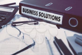 Ring Binder with inscription Business Solutions on Background of Working Table with Office Supplies, Glasses, Reports. Toned Illustration. Business Concept on Blurred Background.