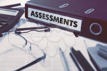 Ring Binder with inscription Assessments on Background of Working Table with Office Supplies, Glasses, Reports. Toned Illustration. Business Concept on Blurred Background.